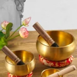 A set of three Brass Buddha Sound Bowls | Nepalese Meditation Singing Bowls with wooden mallets sit on patterned cushions. In the background, pink lotus flowers and a greyscale image of a robed figure with prayer beads appear against a light wall.