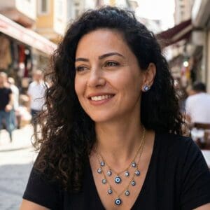 A woman with curly dark hair and a black top smiles while sitting outdoors at a busy street café. She wears matching blue evil eye symbolic jewellery, including a sterling silver pendant. The background is slightly blurred, showing people and shops.