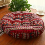 Round red bohemian meditation cushion with geometric patterns placed on a wooden floor near a plant and cup of tea.