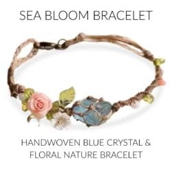 The Sea Bloom Bracelet is a handwoven blue crystal bracelet with a large crystal centerpiece, pink and white flower charms, green leaves, and a pink rose on tan cord—capturing the essence of nature’s beauty.