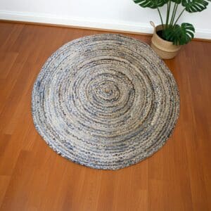 The Round Jute and Recycle Denim Rug - 150 cm in blue, gray, and beige lies on a medium-tone wooden floor. A large-leafed green plant in a woven basket sits in the corner against a white wall.