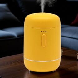 A yellow Essential Oil Waterless Nebuliser with a central button and indicator lights sits on a wooden table, releasing a visible mist. A dark sofa and cushions are softly blurred in the background.