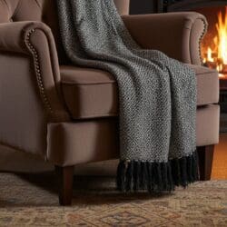 A brown armchair with nailhead trim is draped with a Boho Comfort Throw - Charcoal, featuring fringed edges. The chair sits on a patterned rug near a cozy fireplace, where visible flames glow in the background.