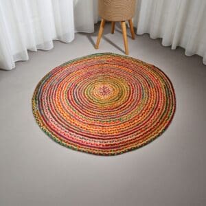 The Round Jute and Recycled Cotton Rug - 150 cm, featuring multicolored braided rings in red, yellow, green, and orange, is displayed on a light gray carpeted floor with white sheer curtains and a wooden tripod table nearby.