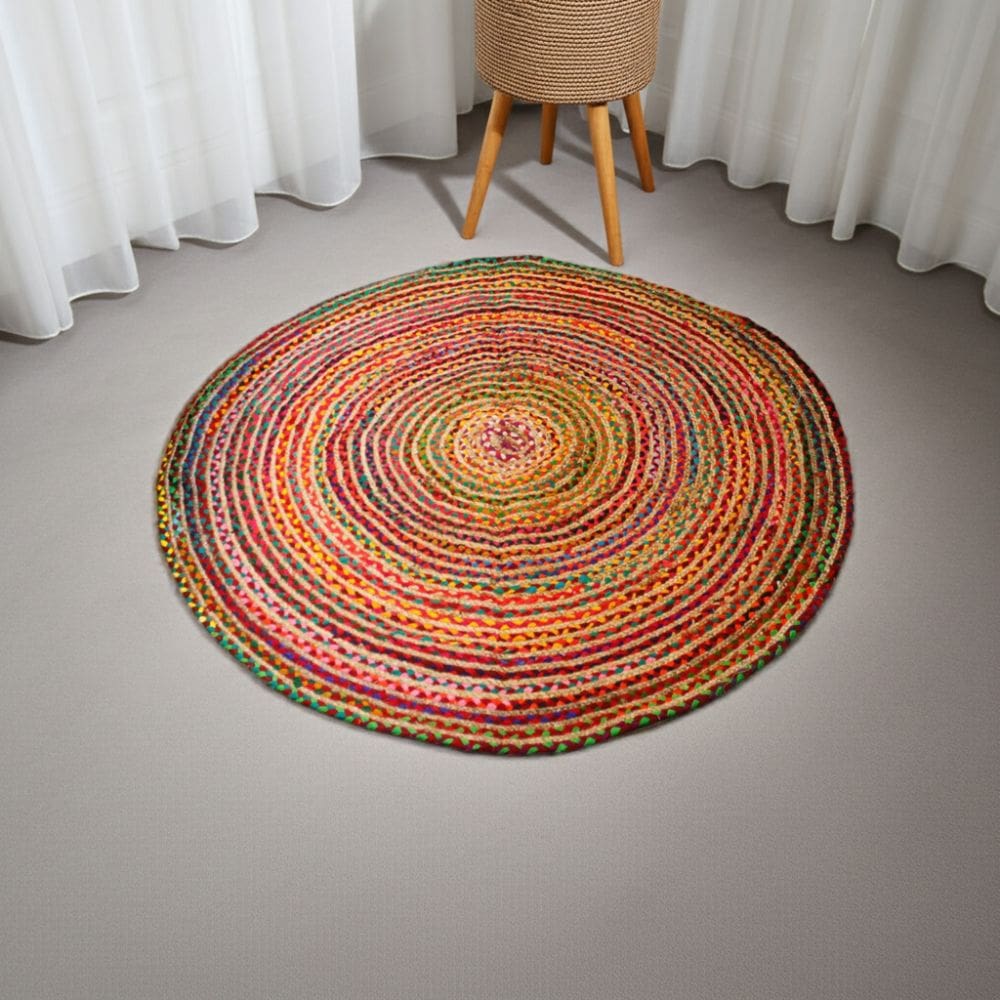 The Round Jute and Recycled Cotton Rug - 150 cm, featuring multicolored braided rings in red, yellow, green, and orange, is displayed on a light gray carpeted floor with white sheer curtains and a wooden tripod table nearby.