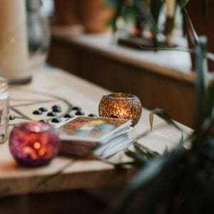A wooden table holds two mosaic glass candle holders, one orange and one purple, both glowing. A deck of tarot cards and black stones suggest a space where one might wonder, is manifesting real? Green leafy plants and a window glow in the soft light.