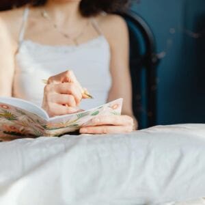 A woman in a white camisole sits on a bed, journaling in a floral-patterned notebook and reflecting on manifesting mistakes. She is partially visible from the shoulders down, with soft natural light highlighting the pillow and bedspread in the foreground.
