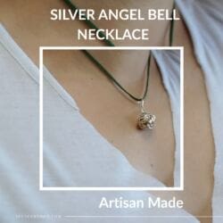 A close-up of a person wearing the Silver Angel Bell Necklace 23mm Artisan Made on a dark cord against bare skin and a white V-neck shirt. “Silver Angel Bell Necklace 23mm” appears at the top, “Artisan Made” at the bottom.