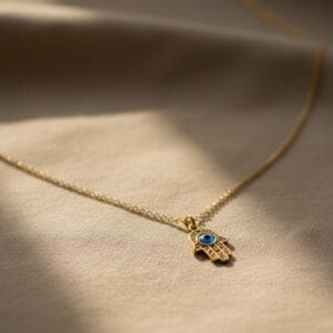 A delicate gold necklace with a Hamsa hand pendant featuring a vibrant hamsa Turkish eye in the center rests on beige fabric, softly lit by natural light. The finely detailed chain and pendant create a subtle, elegant look.