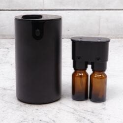 The Double Switchable Waterless Nebuliser – USB-C Essential Oil Diffuser in sleek black rests on light marble, its detached cartridge holding two amber bottles. Light gray tiles in the background give a clean, minimal look.
