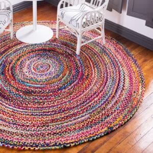 The Round Jute and Recycled Cotton Rug - 150 cm, multicolored and braided, lies on a polished wooden floor in a corner, encircled by two white chairs with striped cushions and a white round table; gray-trimmed windows are in the background.