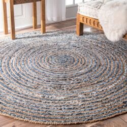The Round Jute and Recycle Denim Rug-90 cm, featuring blue, beige, and white concentric circles, is placed on a wooden floor beneath a chair and bed with a white faux fur throw by a window with sheer curtains.