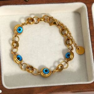 The Lucky Turkish Evil Eye Beads Chain Bracelet for women features gold plating, blue evil eye beads, an adjustable clasp, and a disc charm, displayed in a cream jewelry box. Made from waterproof stainless steel for lasting wear.