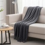 The Good Nite Throw Blanket Fluffy Fleece (127 x 152 cm, Grey) with fringes is draped over a beige sofa. In front, a round wooden coffee table holds a cup and book, surrounded by light curtains, a floor lamp, and cozy neutral decor.