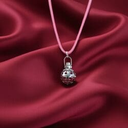 The Sterling Silver Angel Bell Pendant – Romance – 18mm hangs from a pink cord necklace, resting on burgundy satin fabric. This ornate charm features intricate cutouts and patterns, adding an elegant romantic touch.