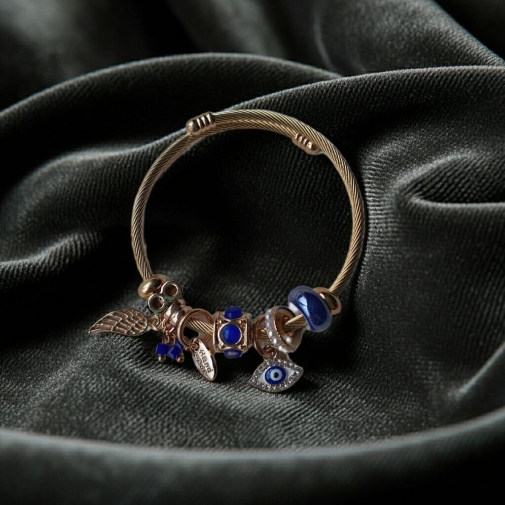 The Gold-Tone Charm Bracelet with Blue Beads & Evil Eye Detail, featuring wing and bead accents, is displayed on softly folded dark green velvet fabric.