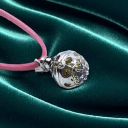 The Sterling Silver Angel Bell Pendant | 16mm Handmade features intricate filigree with gold accents and hangs on a pink suede cord, displayed atop emerald green satin to highlight its elegant openwork design.