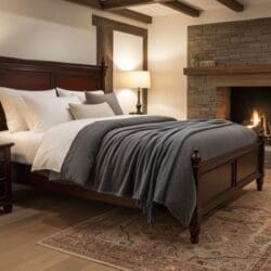 A cozy bedroom with a wooden bed frame, white and gray bedding, the Good Nite Throw Blanket Fluffy Fleece Flannel (127 x 152 cm, Grey), a stone fireplace, patterned rug, bedside tables with lamps, and wooden ceiling beams creates a warm atmosphere.
