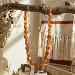 The Amber-Tone Beaded Necklace with Gold Chain Accent hangs on a rustic branch beside dried flowers and a beige woven wall hanging with red stripes, creating a cozy boho vibe. The Spencer Vibes logo appears in the corner.