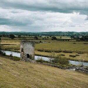 A weathered stone tower ruin stands on a grassy hill overlooking a winding river, offering rest and relevance in the tranquil rural landscape, where fields, sparse trees, and rolling hills stretch beneath a cloudy sky.
