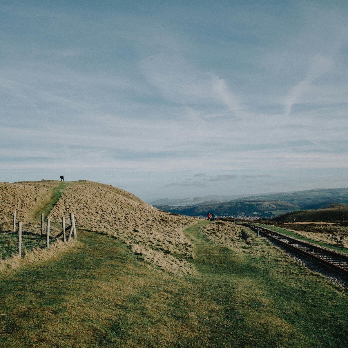 Grassy hills with a narrow path winding alongside a wooden fence and railway tracks reflect the quiet pressure of the landscape. One person stands on a hilltop to the left, while two others walk further along the winding trail under a vast, partly cloudy sky.