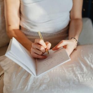 A person in a white sleeveless top writes in cursive with a gold pen, notebook balanced on their lap—where productivity becomes a personality trait, embodied in each elegant stroke.