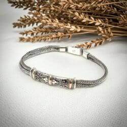 The Silver & Gold Twin Chain Bracelet – Unisex Design, featuring detailed cylindrical accents and a clasp, rests on light fabric. Dried wheat stalks in the background add a rustic, natural touch to the scene.