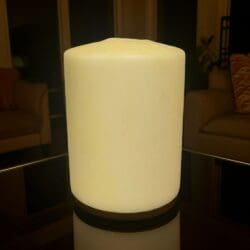 A large, unlit Church Candle – 200 × 150 mm – Three Wick sits on a reflective glass table. In the background are softly lit windows, a plant, and two beige couches.