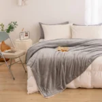 A neatly made bed with white and grey pillows features the Ultra-Soft Sherpa Fleece Blanket in Calm Grey. A book and glasses rest on the cosy blanket, while a round bedside table nearby holds flowers, a clock, and a brown vase on the wooden floor.
