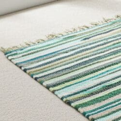 The Large Rag Rug - 150x90cm, premium quality in Natural Greens, with green, blue, white, and grey stripes plus fringe lies on a beige carpet in a bright room, adding a colourful accent to the neutral decor.