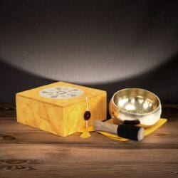 The Chakra Singing Bowl Set – Tibetan Style for Meditation & Sound Healing includes a brass singing bowl on a yellow cloth with a matching box and a wooden mallet, all beautifully arranged on wood against a softly lit background—ideal for sound healing sessions.