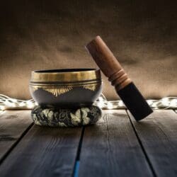 The Sacred Geometry Tibetan Singing Bowl Set – Etched Brass for Sound Healing rests on a patterned cushion with a wooden mallet, glowing fairy lights, and a rustic wooden surface against a dark, textured backdrop.