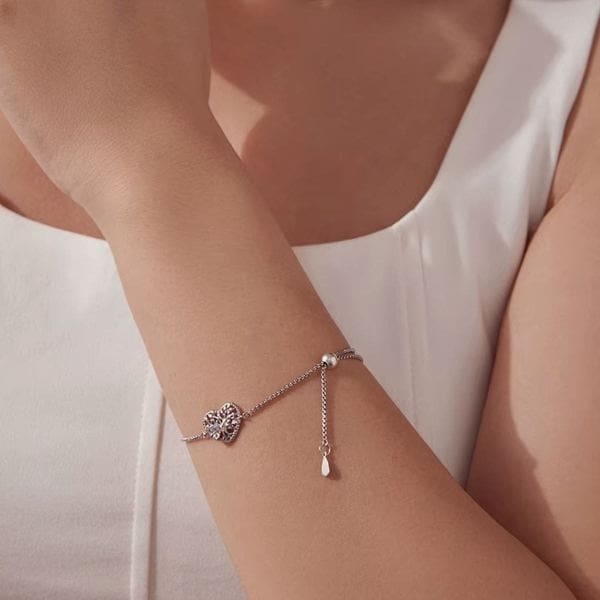 A person in a white sleeveless top showcases the 925 Sterling Silver Tree of Life Bracelet with Moonstone Heart, featuring an adjustable chain and a softly lit background that highlights its elegance.
