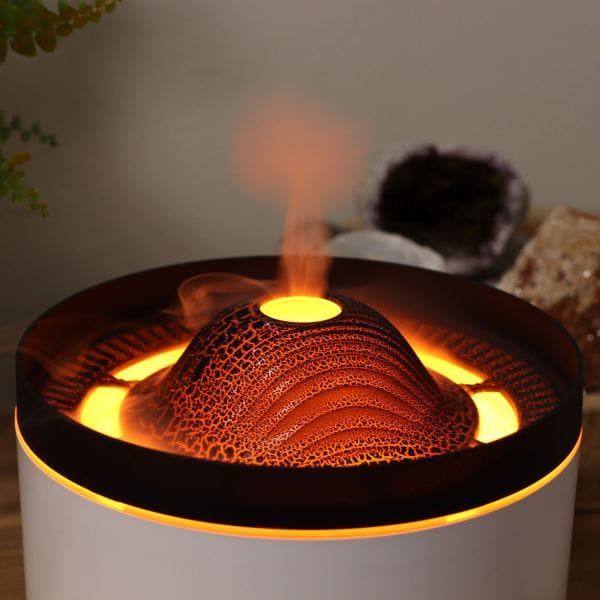 A close-up of the Essential Oil Diffuser – Volcano Effect glows orange and releases mist like lava, creating a cozy vibe. Shown on a white surface with blurred crystals and plants. Includes free Lavender 10ml and 15% off Massage Oil.